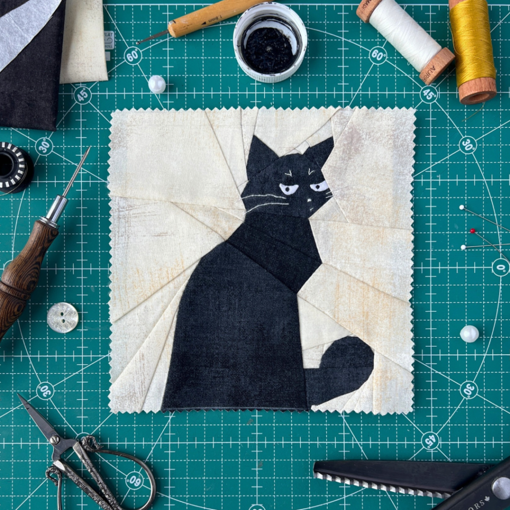 What black cat quilt block pattern displayed on green cutting mat with sewing tools and thread