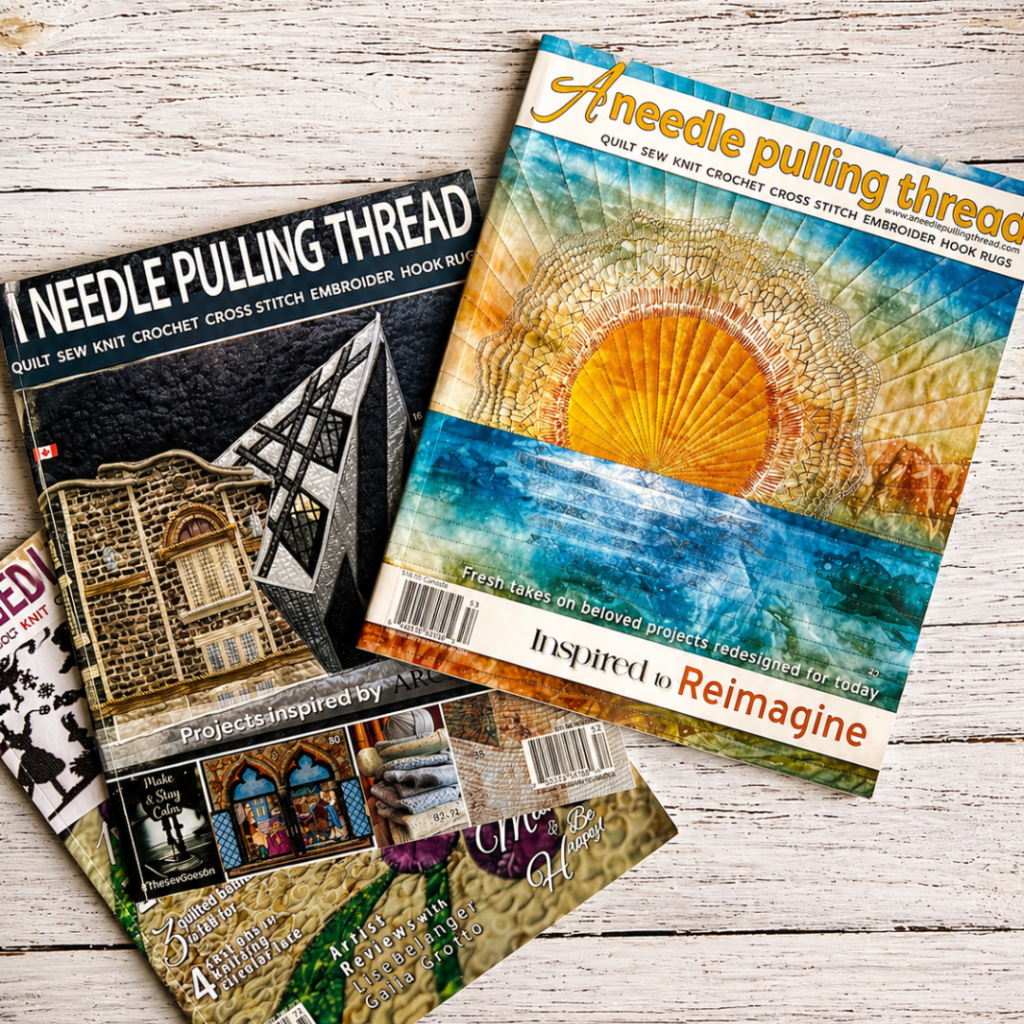 Issue 71 of A Needle Pulling Thread displayed with two earlier magazine issues on white wood background