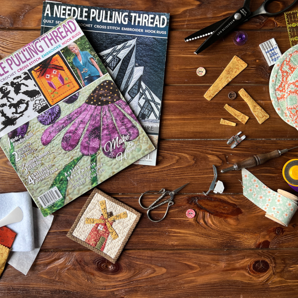 Styled flat lay with quilting tools, magazine issues, fabric pieces, and small windmill block on wooden table