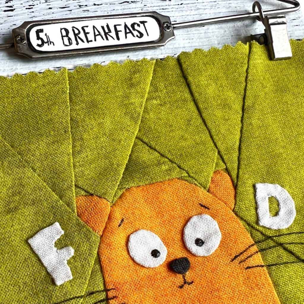 Close-up of 5th Breakfast ginger cat quilt block showing cat face and letter details on green patchwork background