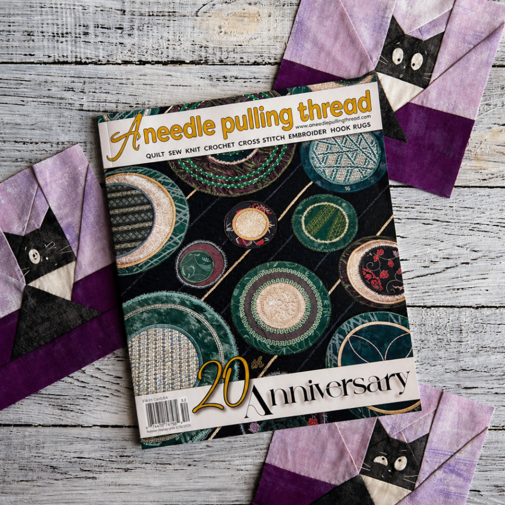 Cover of A Needle Pulling Thread 20th Anniversary issue on white wood background with three cat quilt blocks