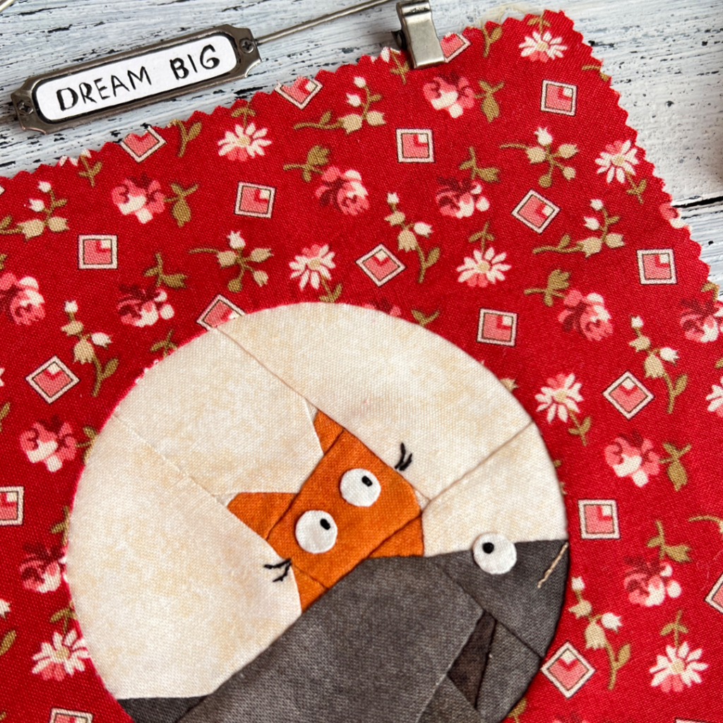 Close-up of Dream Big cat quilt block showing kitty face and large fish detail on red floral background