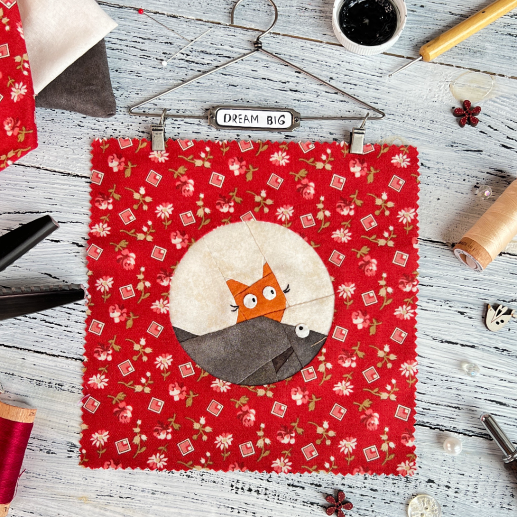 Dream Big cat quilt block pattern featuring a ginger cat peeking over a large fish on red floral background with sewing tools