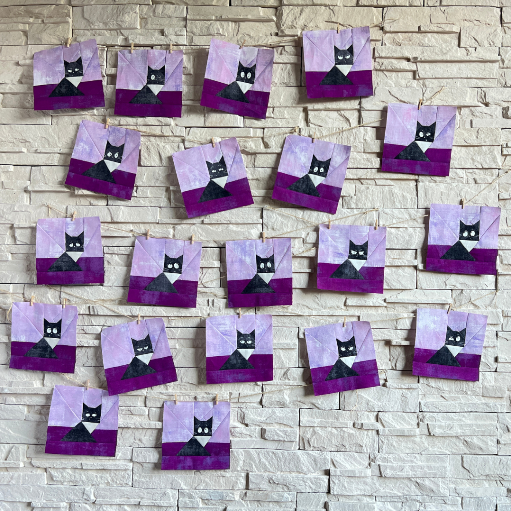 Wall display of twenty Faces of 20 Whiskers black cat quilt blocks in purple and black fabrics