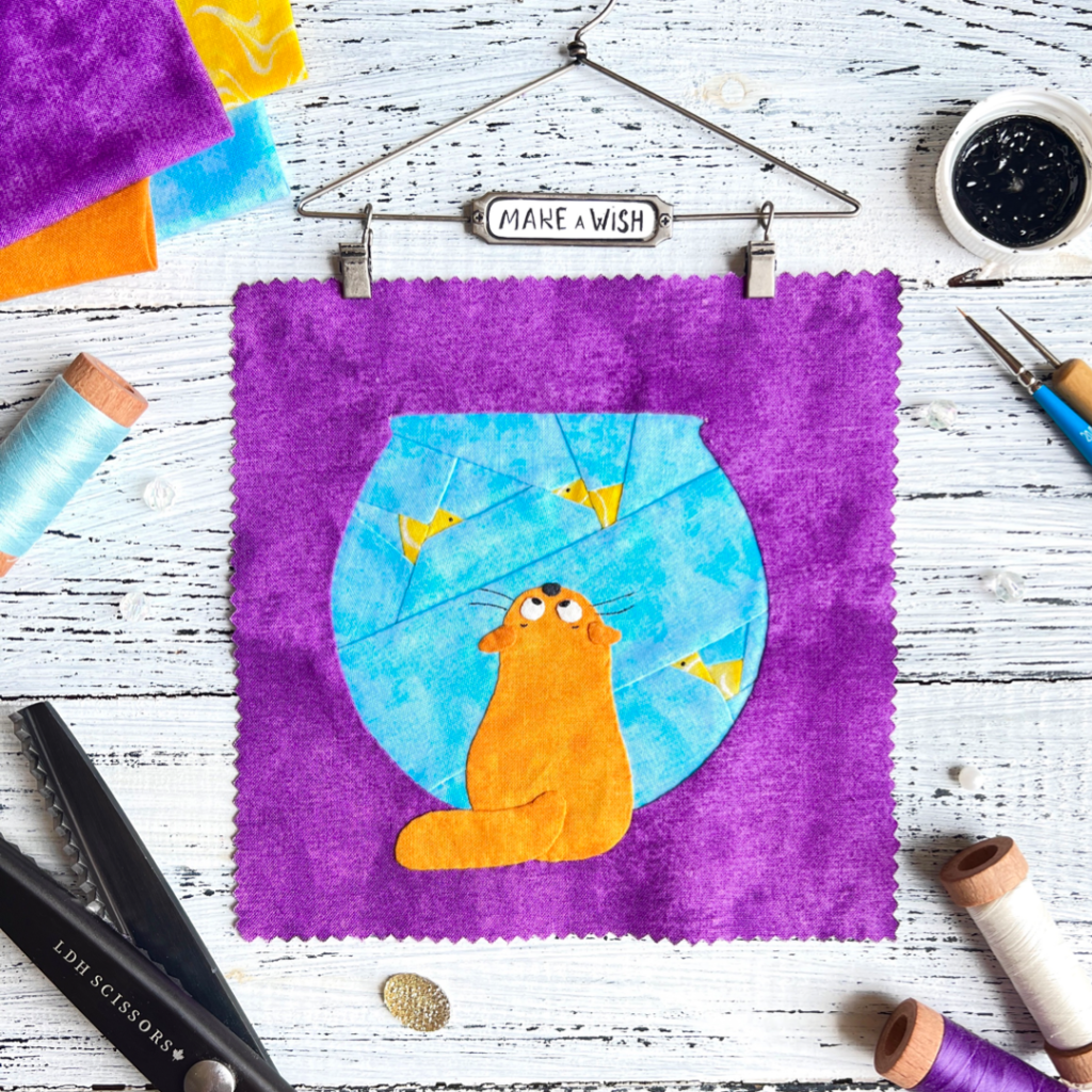 Make a Wish cat quilt block pattern featuring a ginger cat and fishbowl on a purple background with sewing tools
