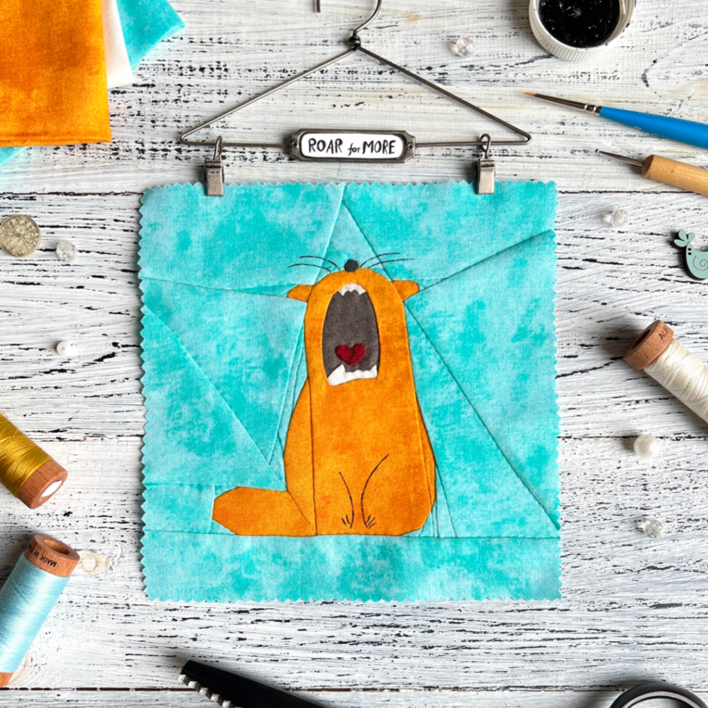 Roar for More ginger cat quilt block pattern on a turquoise background with sewing tools and hanger display